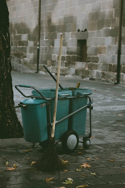 A street scene showing a blue commercial cleaning trolley equipped with various cleaning tools, positioned on a paved sidewalk next to a tree with a broom leaning against it. The trolley contains cleaning buckets and supplies, and the sidewalk surface appears damp, suggesting recent cleaning activity. In the background, there is a stone wall with a small opening, and a few fallen leaves are scattered around. The scene is lit by natural daylight, emphasizing the wet pavement and clean appearance of the trolley, illustrating the typical equipment used in professional cleaning services such as those offered by Kentish Town Carpet Cleaners, aligning with surface cleaning and hygiene maintenance for residential or commercial spaces.