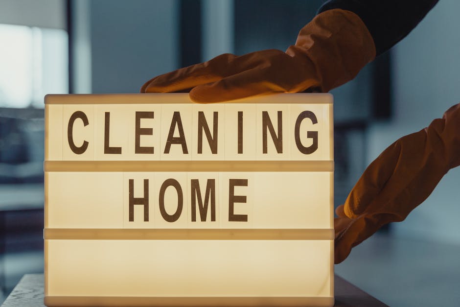 A person wearing brown rubber gloves is arranging a wooden letter lightbox on a table, spelling out 'CLEANING HOME' in black capital letters. The lightbox emits a soft, warm glow, contrasting with the darker, blurred background of an indoor space, possibly a kitchen or living room. The surface of the table appears clean and smooth, with no visible dust or clutter. The image emphasizes the theme of domestic surface cleaning and hygiene, aligning with the services offered by Kentish Town Carpet Cleaners available at the URL https://kentishtowncarpetcleaners.com, to avoid hidden cleaning charges in Kentish Town quotes.