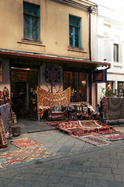 An outdoor shop displays a variety of colorful, patterned rugs and carpets laid out on the pavement and draped over display stands. The shop has an open storefront with textile decorations hanging at 