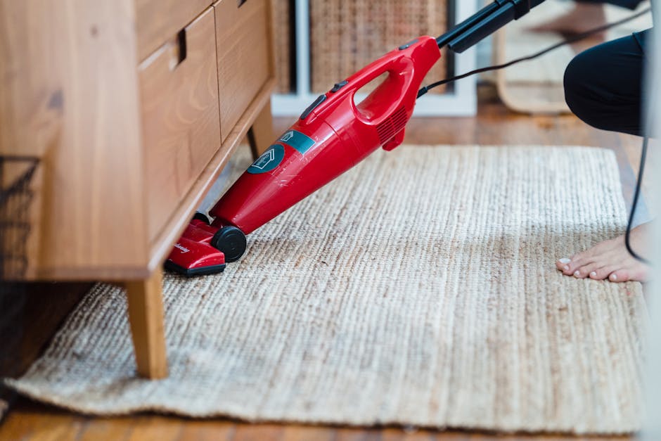 A person vacuuming a beige textured rug with a red handheld vacuum cleaner placed on a wooden floor next to a wooden sideboard. The room is well-lit with natural light, and the furniture appears clean and well-maintained. The vacuum cleaner is turned on, and the person's hand is visible as they guide the device over the rug, demonstrating surface cleaning. The setting suggests a domestic environment focused on maintaining cleanliness and hygiene, with simple and modern decor. Kentish Town Carpet Cleaners emphasise proper cleaning procedures to avoid common mistakes in carpet maintenance, ensuring effective sanitisation and deep cleaning of residential spaces.