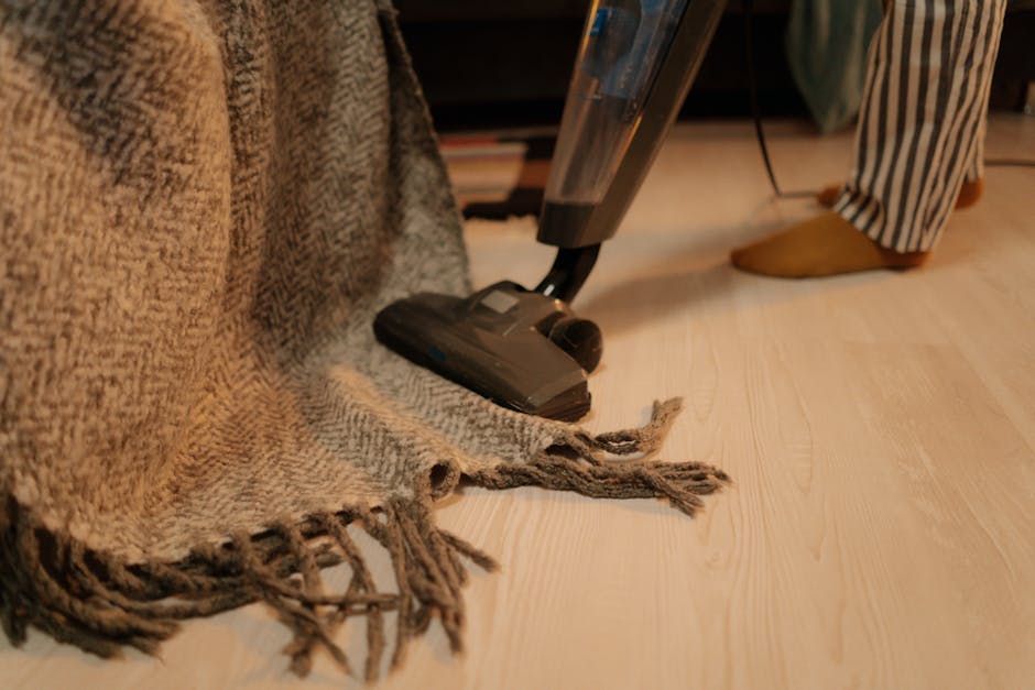 A person vacuuming a beige textured rug with a red handheld vacuum cleaner placed on a wooden floor next to a wooden sideboard. The room is well-lit with natural light, and the furniture appears clean and well-maintained. The vacuum cleaner is turned on, and the person's hand is visible as they guide the device over the rug, demonstrating surface cleaning. The setting suggests a domestic environment focused on maintaining cleanliness and hygiene, with simple and modern decor. Kentish Town Carpet Cleaners emphasise proper cleaning procedures to avoid common mistakes in carpet maintenance, ensuring effective sanitisation and deep cleaning of residential spaces.
