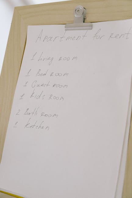 A close-up image of a handwritten list on a white sheet of paper clipped to a wooden board, detailing rooms in an apartment for rent, including a living room, bedroom, guest room, kids' room, bathrooms, and kitchen. The paper appears slightly crumpled, and the handwriting is in pencil, with some words underlined for emphasis. The background suggests an indoor setting with neutral-coloured walls. The focus is on the list, which is not directly related to cleaning or surface surfaces; therefore, no surfaces, materials, or cleaning activity is visible in this image. The image is used on a page related to end of tenancy cleaning for properties on Crowndale Road, with the associated company, Kentish Town Carpet Cleaners, offering professional cleaning services.