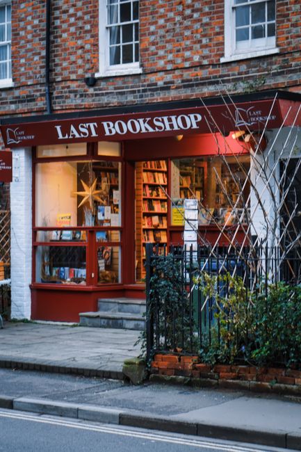 The exterior of the Last Bookshop located in a brick building with white-framed windows, featuring a brown awning with white lettering. The shop window displays shelves filled with books and a decorative star-shaped light. In front of the shop, there is a small paved area with steps leading into the store, and a black metal fence surrounds some greenery and small plants. The scene is lit by warm indoor lighting that highlights the books and interior, suggesting a cozy, inviting atmosphere. This image captures the avenue outside the shop, emphasizing the urban environment typical of Kentish Town, with a focus on the quaint and tidy presentation of the store, which aligns with quality cleaning and maintenance standards.