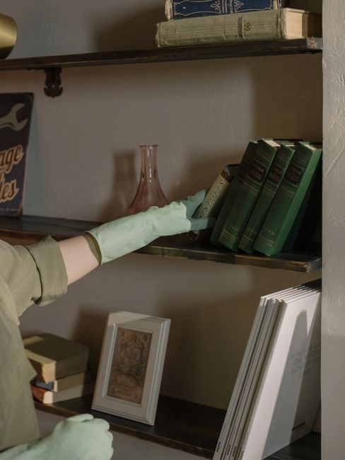 A person wearing light-colored gloves is cleaning a wooden bookshelf in a domestic setting, possibly in a living room or study. The person is reaching out with their right hand to straighten or dust a collection of green hardcover books on the middle shelf. The shelf contains a variety of items, including a pink glass vase, a small framed picture, and a stack of books. The room is lit with soft, natural or ambient lighting, highlighting the surfaces' cleanliness and shine. The shelving unit appears aged with visible wood grain, and the surrounding wall is painted in a neutral, light color. This image illustrates routine surface cleaning and maintenance as part of a professional service by Kentish Town Carpet Cleaners, reflecting deep cleaning and sanitisation practices for a tidy, well-maintained home environment.