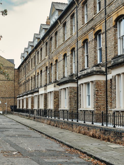 Image of a row of Victorian-style brick residential buildings with multiple stories, featuring white-framed windows and black wrought iron railings along the small front yards. The buildings are bathed in natural daylight, and the pavement in the foreground is clean with some fallen leaves, indicating a well-maintained street. The scene reflects an urban residential area typical of Kentish Town West, where professional cleaning services by Kentish Town Carpet Cleaners may be used for surface cleaning and property maintenance. The overall environment appears tidy and well-kept, emphasizing cleanliness and proper upkeep of the exterior surfaces.