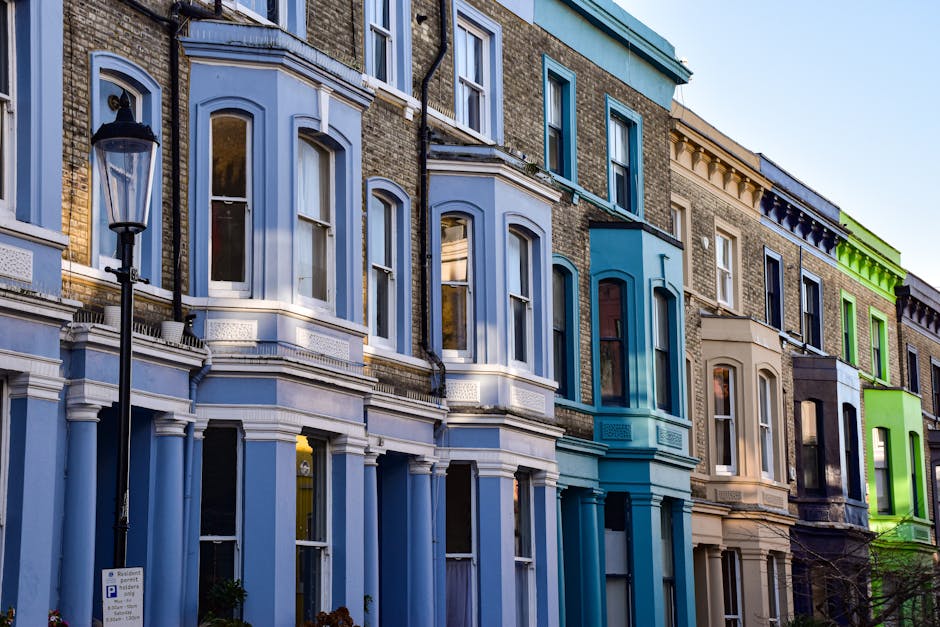 Exterior view of a row of colorful Victorian-style terraced houses in Kentish Town, featuring high-rise bay windows with white trim and brick facades painted in shades of blue, beige, and green. The buildings are illuminated by natural daylight, highlighting the clean and well-maintained surfaces. A classic black street lamp stands in the foreground on the left side, and a parking sign is visible near the sidewalk. The scene captures the charming urban residential architecture typical of the area, with no visible signs of dirt or clutter, emphasizing the neatness and aesthetic appeal of these homes, consistent with the high standards of professional cleaning services like those offered by Kentish Town Carpet Cleaners.