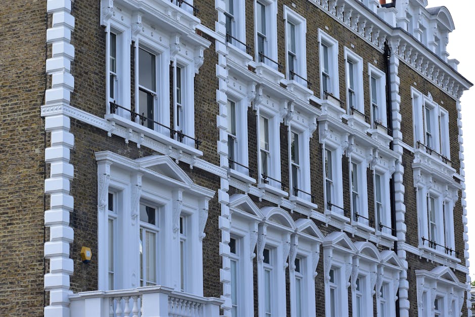Image of a row of Victorian-style brick residential buildings with multiple stories, featuring white-framed windows and black wrought iron railings along the small front yards. The buildings are bathed in natural daylight, and the pavement in the foreground is clean with some fallen leaves, indicating a well-maintained street. The scene reflects an urban residential area typical of Kentish Town West, where professional cleaning services by Kentish Town Carpet Cleaners may be used for surface cleaning and property maintenance. The overall environment appears tidy and well-kept, emphasizing cleanliness and proper upkeep of the exterior surfaces.