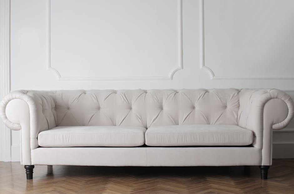 A light beige, tufted fabric sofa with rolled arms and dark wooden legs situated against a white wall with decorative paneling. The surface of the sofa appears clean and well-maintained, demonstrating a smooth, dust-free upholstery. The room has soft, natural lighting that highlights the pristine condition of the furniture. The flooring is parquet wood, adding to the elegant and tidy appearance of the space. This setting exemplifies professional domestic cleaning, emphasizing surface cleaning and furniture sanitisation, as provided by Kentish Town Carpet Cleaners, near Kentish Town station NW5.