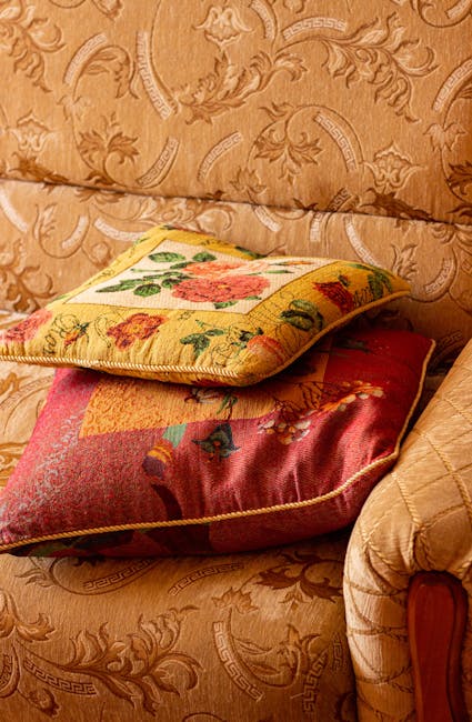 Close-up of a traditional upholstered sofa with a floral patterned fabric in warm beige tones, featuring intricate scroll and leaf motifs. Resting atop the sofa are two decorative cushions with vibrant, embroidered floral designs in shades of red, pink, green, and yellow, adding a splash of color. The cushions have a soft, plush appearance and are positioned slightly askew. The scene is illuminated by natural or soft indoor lighting, highlighting the textures and patterns of the fabric and cushions, conveying a clean and well-maintained domestic setting. This image reflects surface cleaning and hygiene in an interior living space, suitable for content about upholstery cleaning near Kentish Town station NW5, as promoted by Kentish Town Carpet Cleaners.